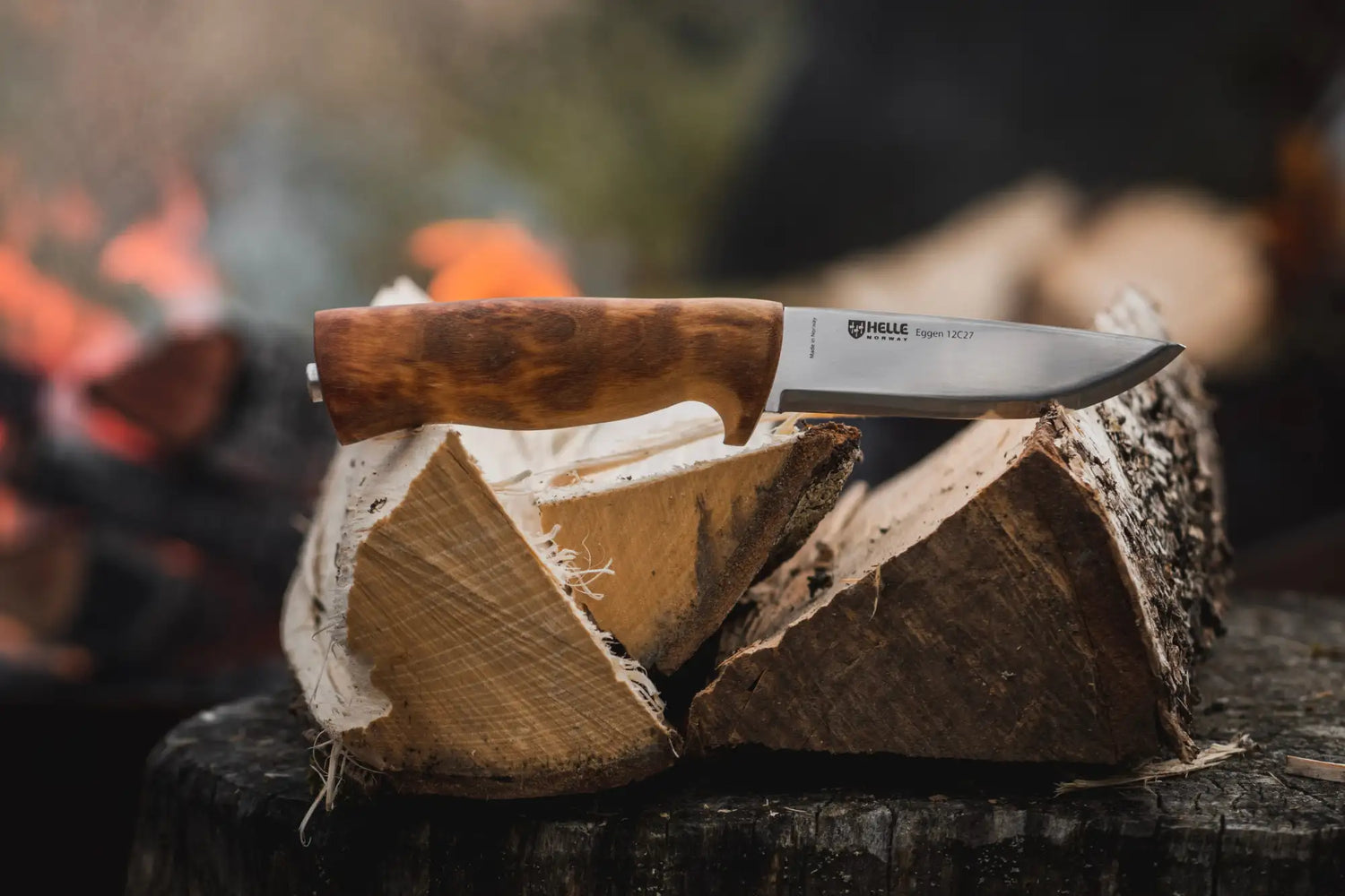 Un couteau helle säppi 302 avec une lame en argent polie et un manche en bois riche, aux tons chauds, reposant sur des bûches fendue.