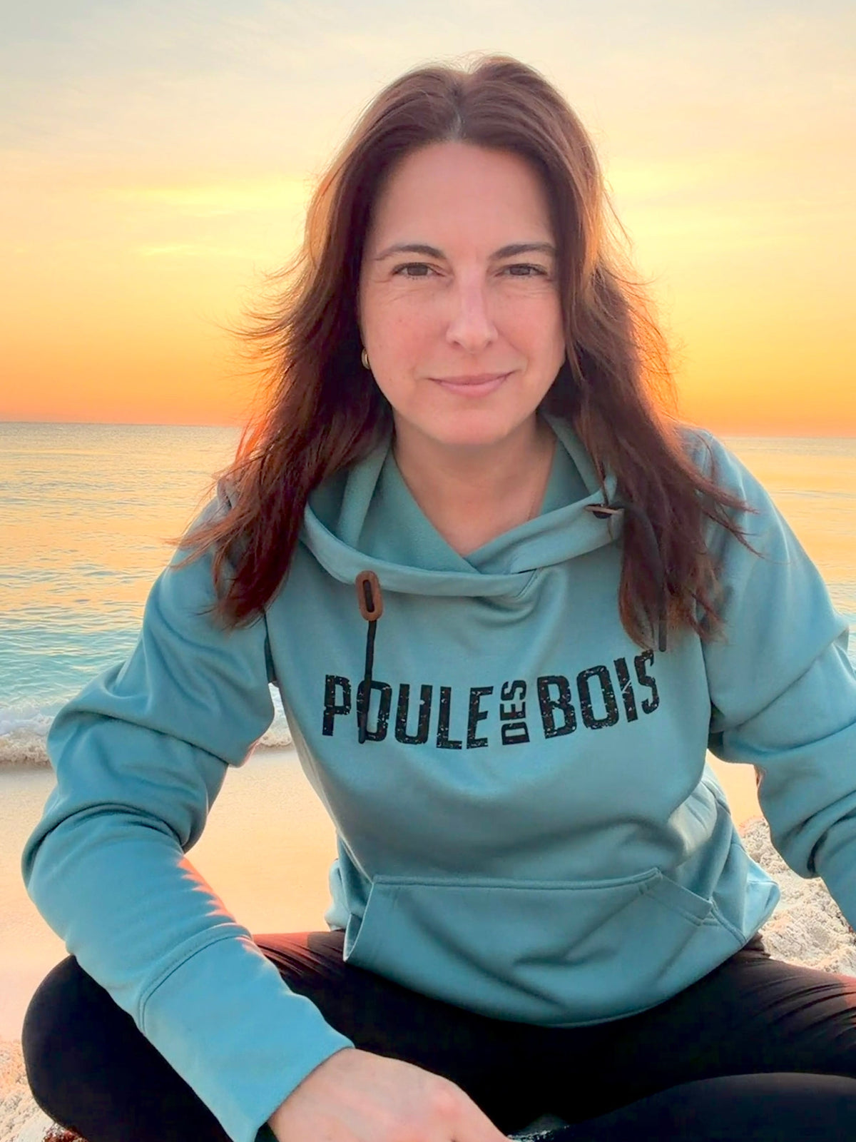 Vue de face du hoodie Yuma sarcelle porté au bord de l’eau, logo noir sur la poitrine