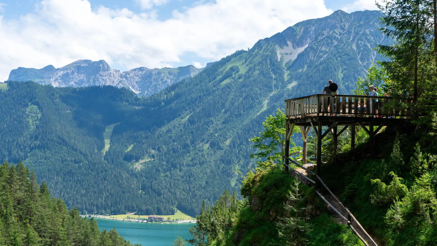 Un ponton d’observation en bois avec une rambarde, perché sur le flanc d’une falaise surplombant un lac turquoise et de hautes montagnes vertes.